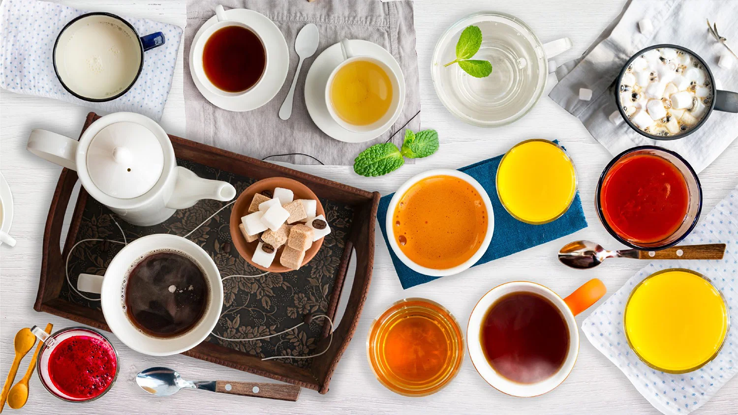 Assortment of colorful hot beverages including coffee, black tea, herbal tea, and hot chocolate in ceramic cups with sugar cubes and teapot on a tray.