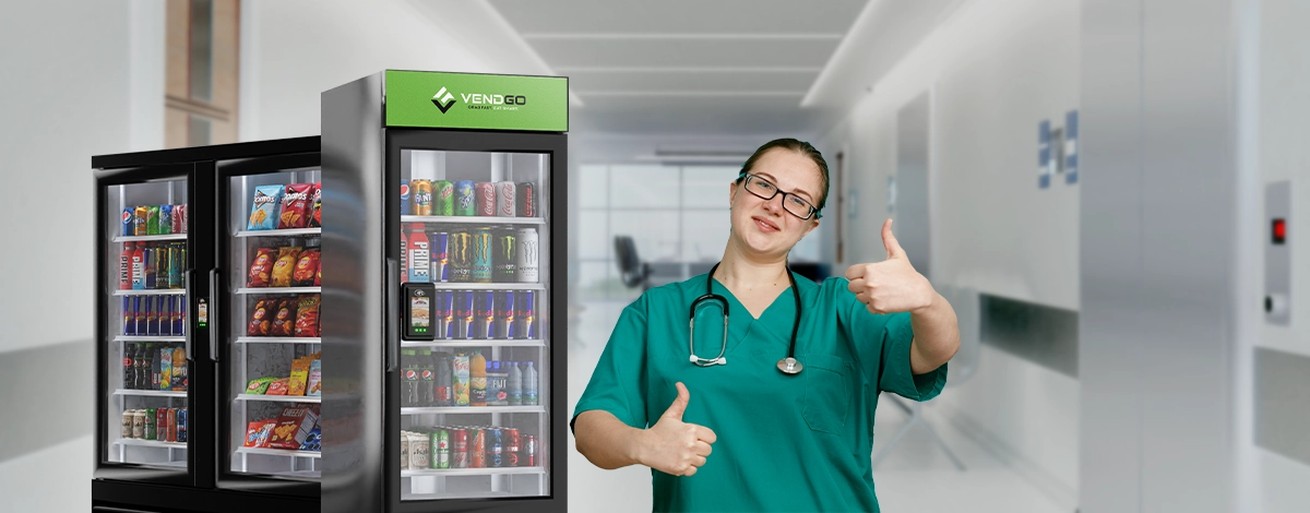 Female nurse in green scrubs giving a thumbs up next to VendGo micro market vending machines in a hospital hallway.