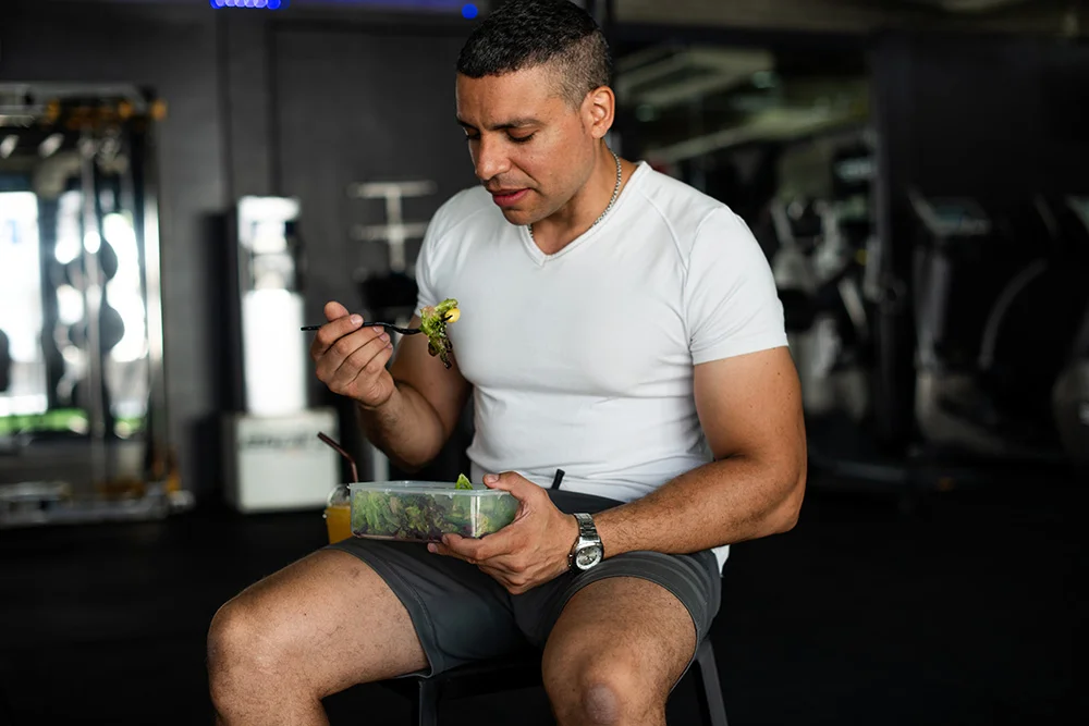 Man in athletic wear sitting on a bench in a gym eating a healthy salad from a container.