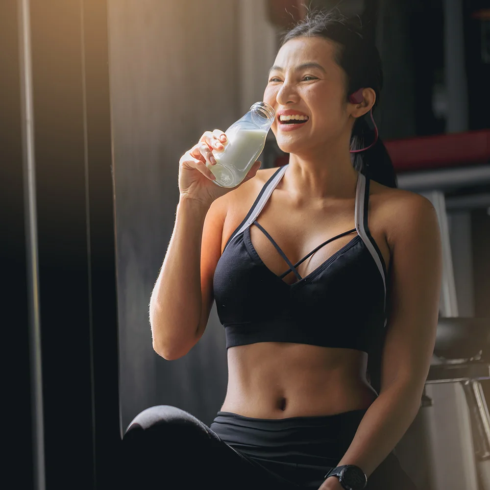 Young woman in sports attire smiling while drinking a bottle of milk or smoothie in a gym after workout.