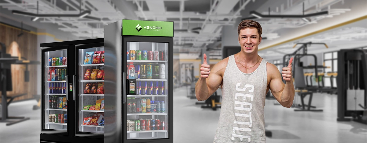 Fit young man giving thumbs up in a gym with Vendgo smart coolers and vending machines stocked with drinks and snacks behind him.