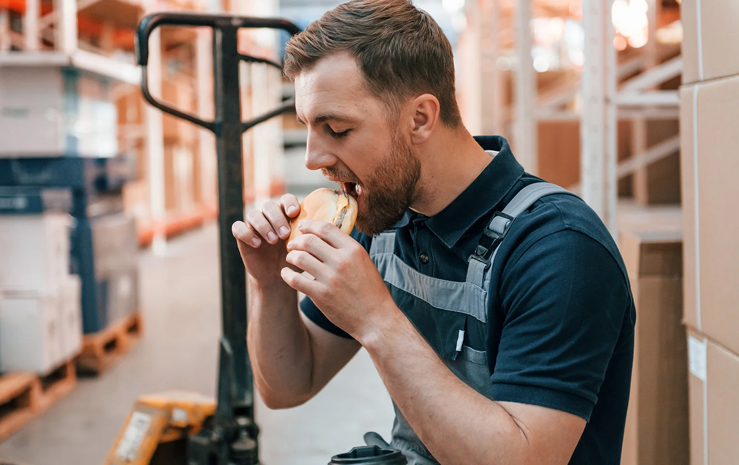 Warehouse employee in uniform taking a lunch break and eating a sandwich among boxes and shelves.