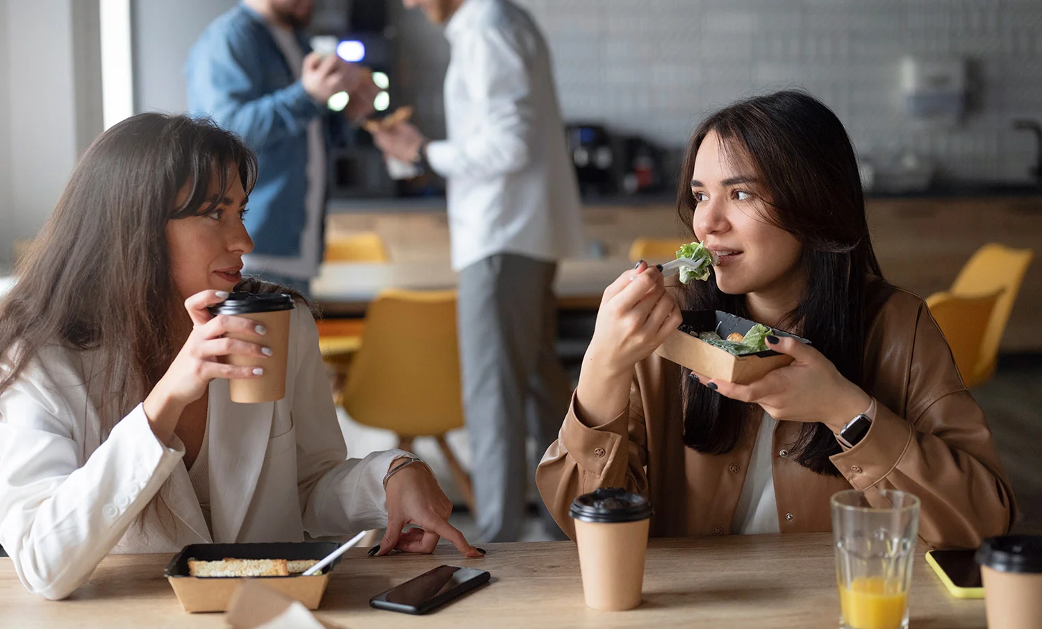 Two female coworkers enjoying a break in a workplace breakroom, one sipping coffee and the other eating a fresh salad from Vendgo's micro market. Colleagues and vending area blurred in the background, representing healthy vending and smart cooler services in Atlanta.