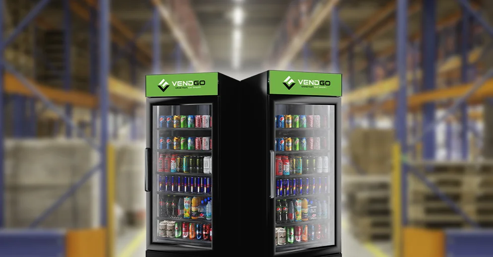 Two Vendgo vending coolers stocked with various drinks positioned inside a warehouse, with blurred shelves and pallets in the background.