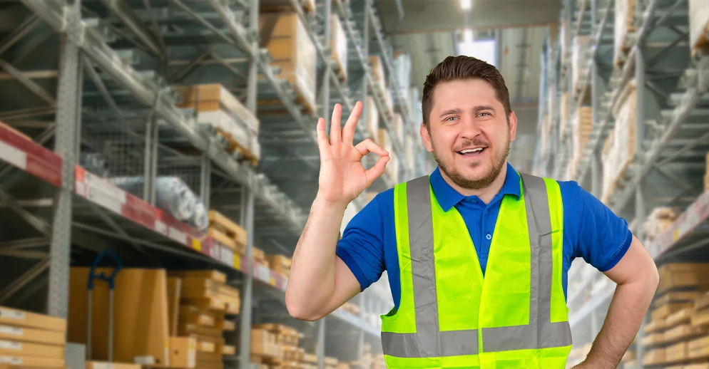 Smiling warehouse worker wearing a high-visibility safety vest standing in an aisle with shelves of boxes, giving an OK hand gesture at a Vendgo-serviced logistics center in Atlanta.