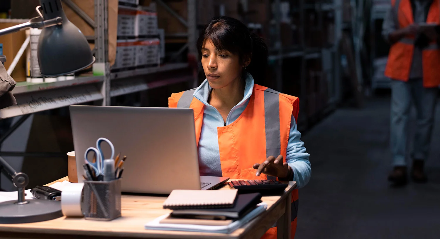 Young woman wearing a reflective safety vest working on a laptop at a desk inside a warehouse with shelves and boxes. She represents Vendgo's logistics team managing micro market vending and smart cooler services for manufacturing facilities in Atlanta.